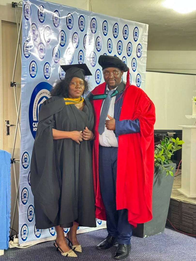 A graduate with a faculty member in front of the Caring Hearts banner